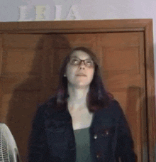 a woman wearing glasses and a denim jacket is standing in front of a door with the word leia on it .