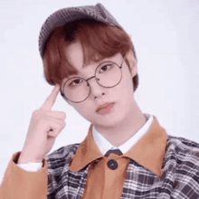 a young man wearing glasses , a hat and a plaid jacket is pointing at his forehead .
