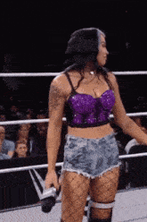 a woman in a purple crop top and shorts is standing in a wrestling ring holding a microphone .