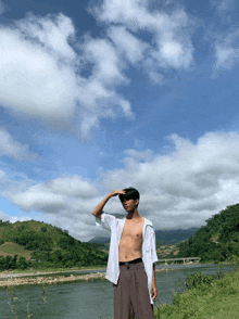 a shirtless man stands in front of a body of water with mountains in the background