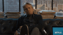 a man sits on a couch in front of a stack of newspapers and a netflix screen