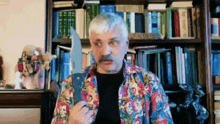 a man with a mustache is holding a large knife in front of a bookshelf