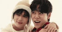 two young men are standing next to each other and hugging each other . one of the men is wearing a hat .