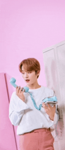 a young man in a white shirt and pink pants is holding a blue telephone in front of a pink wall .
