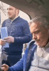 a man wearing headphones is sitting in a car with another man holding a piece of paper .