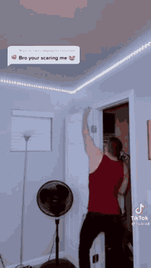 a man in a red tank top is standing in front of a door with a fan in a room .
