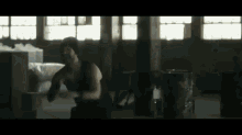 a man in a black tank top is squatting in a dark room with a pepsi barrel in the background