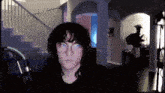 a woman wearing glasses looks at the camera in a dark room with stairs in the background