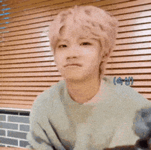 a young man with pink hair is making a funny face while sitting in front of a window with blinds .