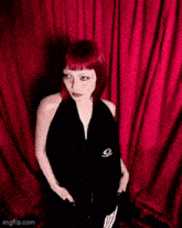 a woman with red hair is standing in front of a red curtain with her hands in her pockets
