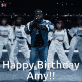a man in a blue jacket stands in front of a group of dancers and says " happy birthday amy !!! "