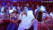 a man in a white robe sits in front of a crowd of people with kvvcsr written on the bottom right