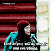 a woman in a red hat is standing in front of a red wall and says `` look at you , still all muscle and everything ''