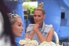 two women are sitting at a table with flowers in front of them and one is crying