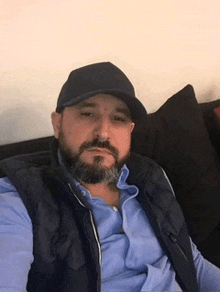 a man with a beard is sitting on a couch wearing a hat and a vest .