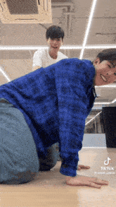 a man in a blue plaid shirt is kneeling down on the floor while another man stands behind him .