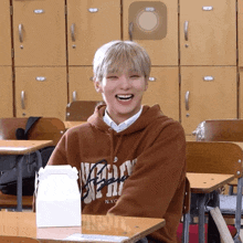 a boy wearing a brown nyc hoodie sits at a desk in a classroom