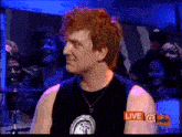 a man with red hair is wearing a black tank top and a necklace while standing in front of a crowd .