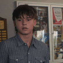 a young man in a striped shirt stands in front of a bulletin board with a poster that says donate blood