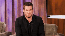 a man in a suit and black shirt is sitting in a chair making a funny face .
