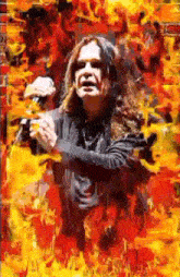 a man with long hair is standing in front of a wall of fire holding a microphone