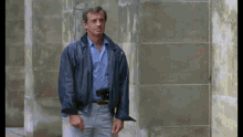 a man wearing a blue shirt and a blue jacket stands in front of a stone wall