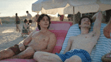 two men are laying on a beach under an umbrella and one has the number 8 on his shorts