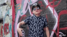 a young man wearing sunglasses and a baseball cap is standing in front of a graffiti covered wall .