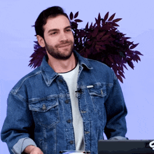 a man in a denim jacket stands in front of a laptop and a plant