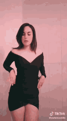 a woman in a short black dress is dancing in front of a pink wall with tiktok written on the bottom