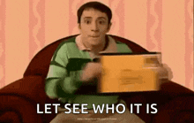 a man in a blue shirt is sitting in a chair holding a box and saying `` let see who it is `` .