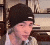 a man wearing a black beanie is sitting in front of a bookshelf and making a funny face .