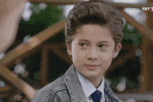 a young boy in a denim jacket and tie looks at the camera with a trt logo in the corner