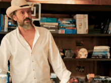 a man in a white shirt and cowboy hat stands in front of a bookshelf with kanald.com.tr written in the corner