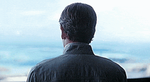a man in a denim jacket looks out a window at the ocean