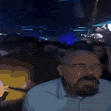 a man with a beard and glasses is sitting in front of a crowd in a dark room .
