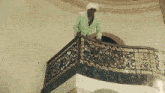 a man in a green jacket is standing on a balcony with a wrought iron railing .