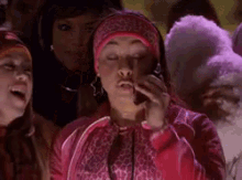 a woman in a pink jacket is talking on a cell phone while standing in front of a crowd of people .