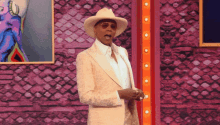 a man in a white suit and cowboy hat is standing in front of a brick wall .