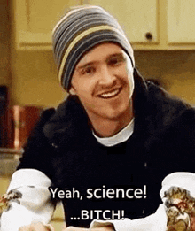 a man wearing a beanie is sitting at a table holding a sandwich and smiling while talking about science .