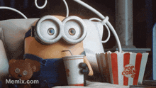 a minion sitting on a bed with a cup of soda and a bucket of popcorn
