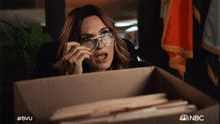 a woman wearing glasses looks out of a cardboard box with the nbc logo on the bottom right