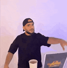 a man with a beard is standing in front of a table with a cup of coffee and a laptop .