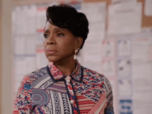 a woman in a colorful shirt and pearl earrings is standing in front of a wall with papers on it .