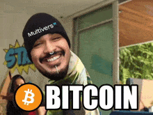 a man wearing a beanie that says multiversx smiles in front of a bitcoin sign