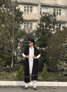 a man wearing a black shawl and white pants stands in front of a building