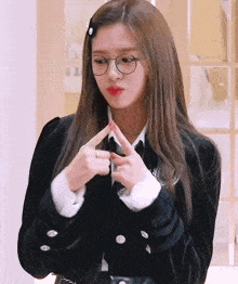 a woman wearing glasses and a black jacket is making a heart shape with her hands