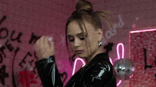 a woman in a black leather jacket stands in front of a wall with graffiti on it that says " love my self "