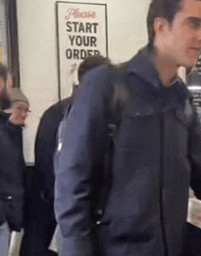 a man in a blue jacket is standing in front of a sign that says please start your order