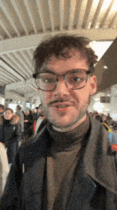 a man wearing glasses and a turtleneck stands in front of a crowd of people